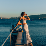 Crop Top and Maxi Skirt set in Blue and White Stripe 100% cotton seersucker deadstock fabric  worn by woman with long brown hair in a bun on a long pier surrounded by blue water, mountains and boats.  She has one hand up to her face touching her sunglasses and looking out towards the sun setting.