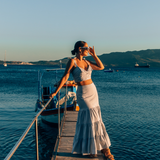 Crop Top and Maxi Skirt set in Blue and White Stripe 100% cotton seersucker deadstock fabric  worn by woman with long brown hair in a bun on a long pier surrounded by blue water, mountains and boats.  She has one hand up to her face touching her sunglasses and looking out towards the sun setting.