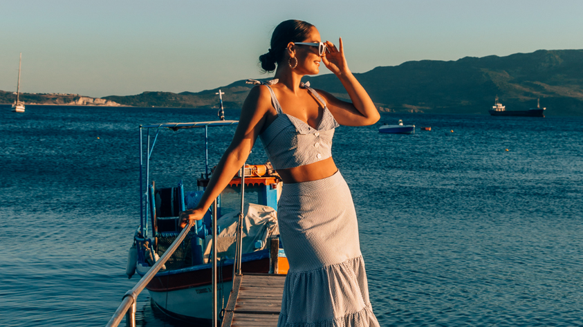 Crop Top and Maxi Skirt set in Blue and White Stripe 100% cotton seersucker deadstock fabric  worn by woman with long brown hair in a bun on a long pier surrounded by blue water, mountains and boats.  She has one hand up to her face touching her sunglasses and looking out towards the sun setting.