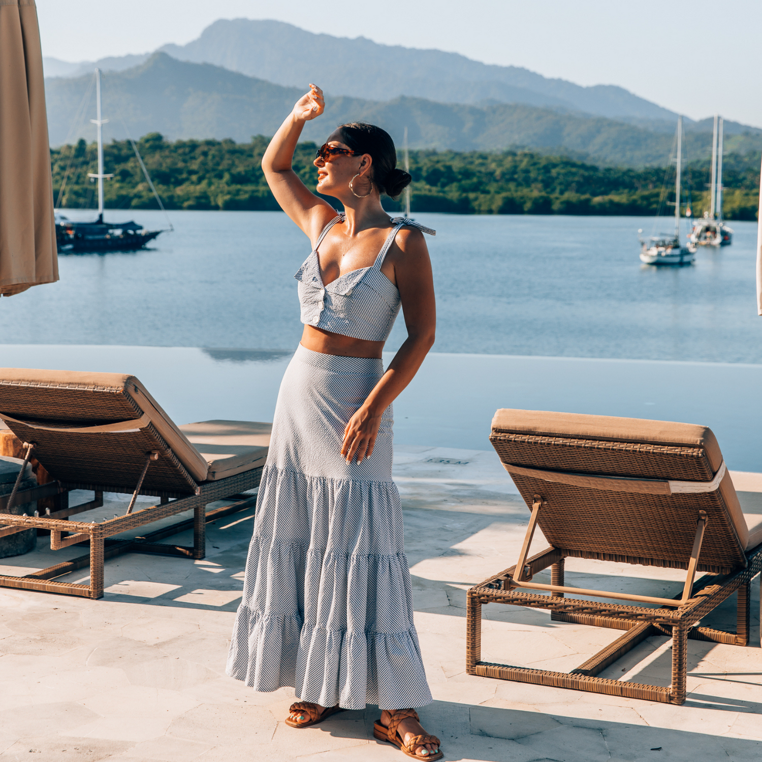 Woman standing in front of infinity pool with deck chairs and ocean in the background dotted with sailing boats and framed by green hill and mountains.  She is wearing a tiered maxi skirt and crop top in blue and white stripes, she has a hand raised to block the sun from her eyes