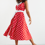 Women standing in front of a white wall , she is facing on a slight angle and looking down over her shoulder towards her outstretched hand.  Her dark curly hair is loosely flowing over her shoulders. She is wearing a red and white polka dot midi dress, which she has twirled in and we can see the voluminous skirt of the dress