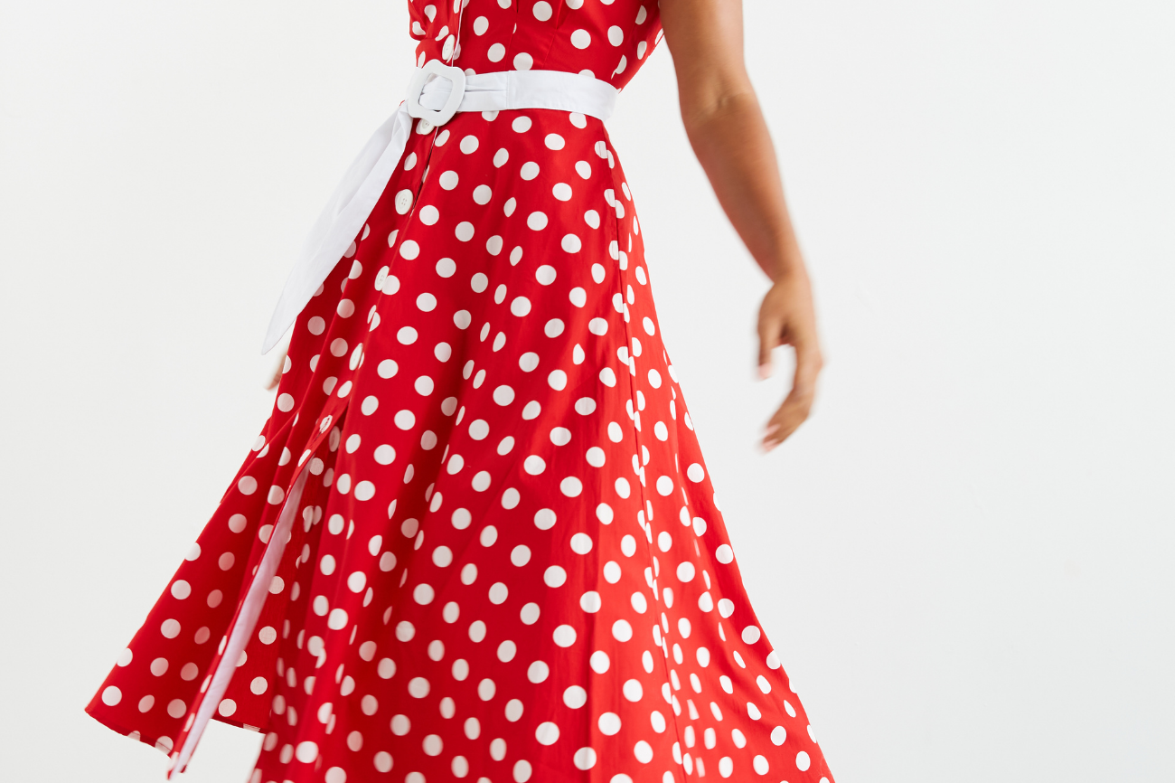 Women standing in front of a white wall , she is facing on a slight angle and looking down over her shoulder towards her outstretched hand.  Her dark curly hair is loosely flowing over her shoulders. She is wearing a red and white polka dot midi dress, which she has twirled in and we can see the voluminous skirt of the dress