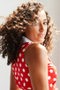 Women standing beside a window , she is facing sideways and looking over shoulder with dark curly hair blowing. She is wearing a red and white polka dot midi dress