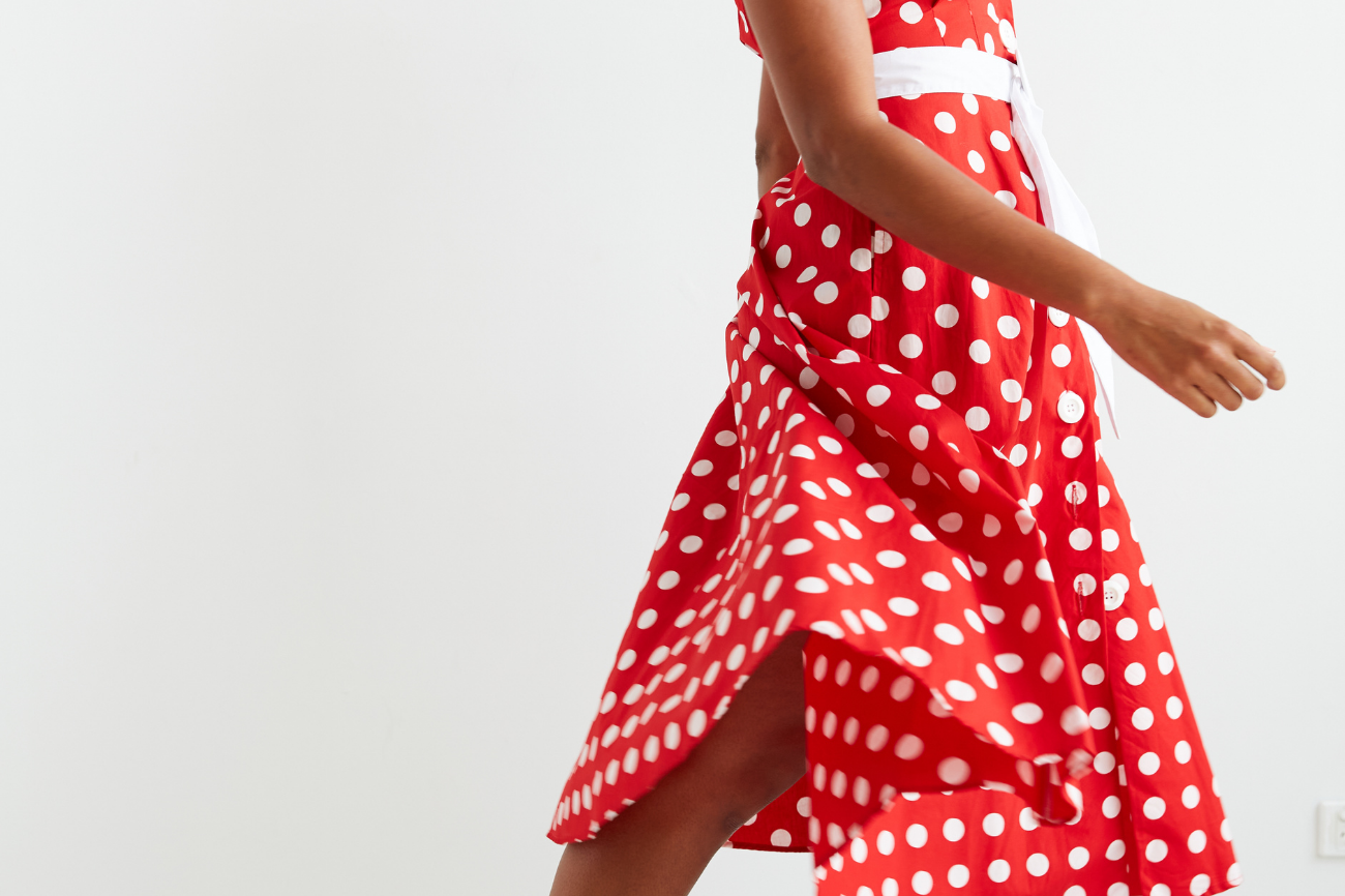 Women standing in front of a white wall , she is facing on a slight angle and looking directly over her shoulder towards the viewer.  Her dark curly hair is loosely flowing over her shoulders. She is wearing a red and white polka dot midi dress, which she has twirled in and we can see the voluminous skirt of the dress