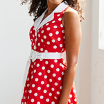 Women standing beside a window , she is facing sideways and looking over shoulder with dark curly hair blowing. She is wearing a red and white polka dot midi dress, image is cropped so you can only see 3/4 of the dress
