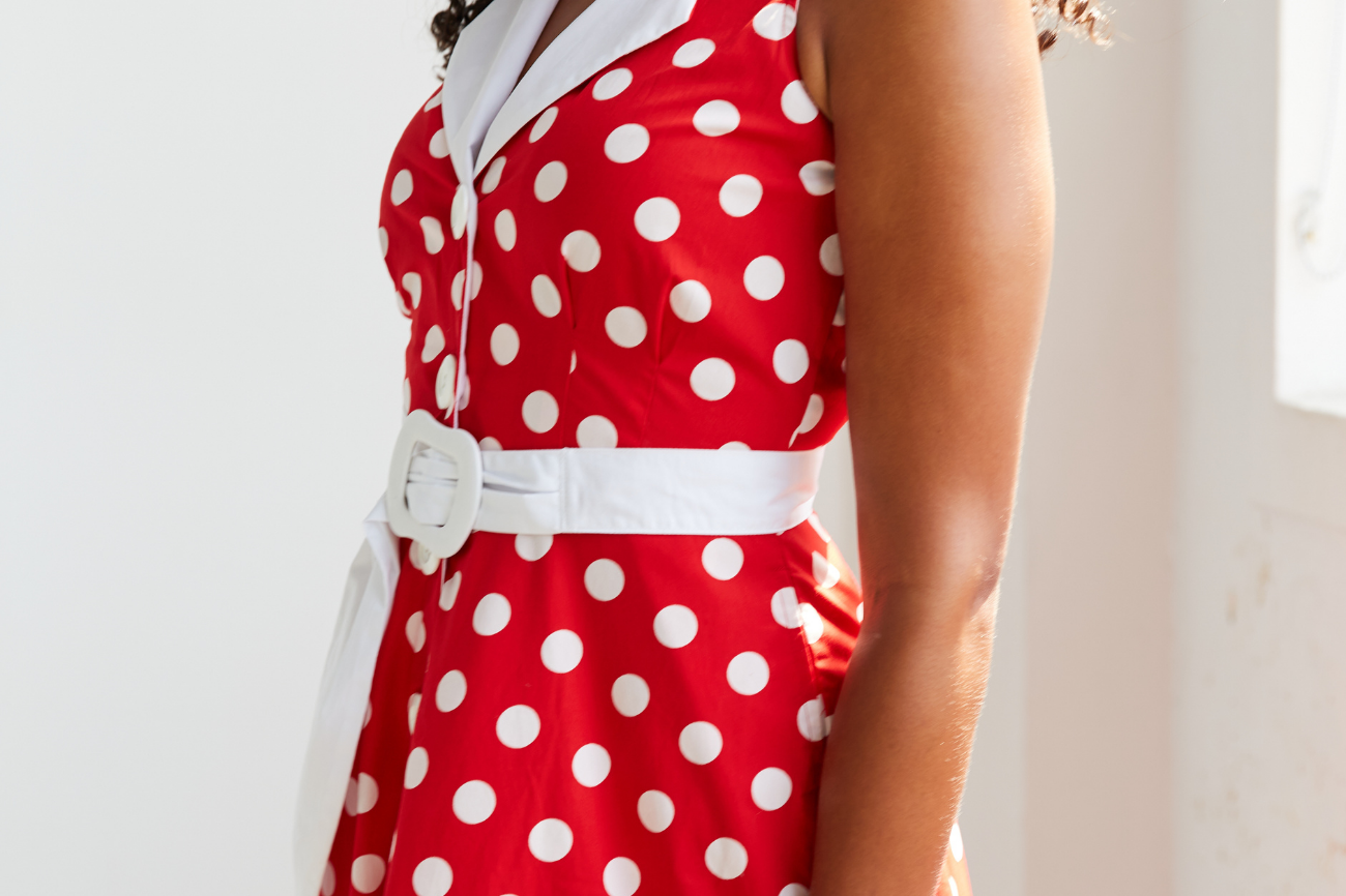 Women standing beside a window , she is facing sideways and looking over shoulder with dark curly hair blowing. She is wearing a red and white polka dot midi dress, image is cropped so you can only see 3/4 of the dress