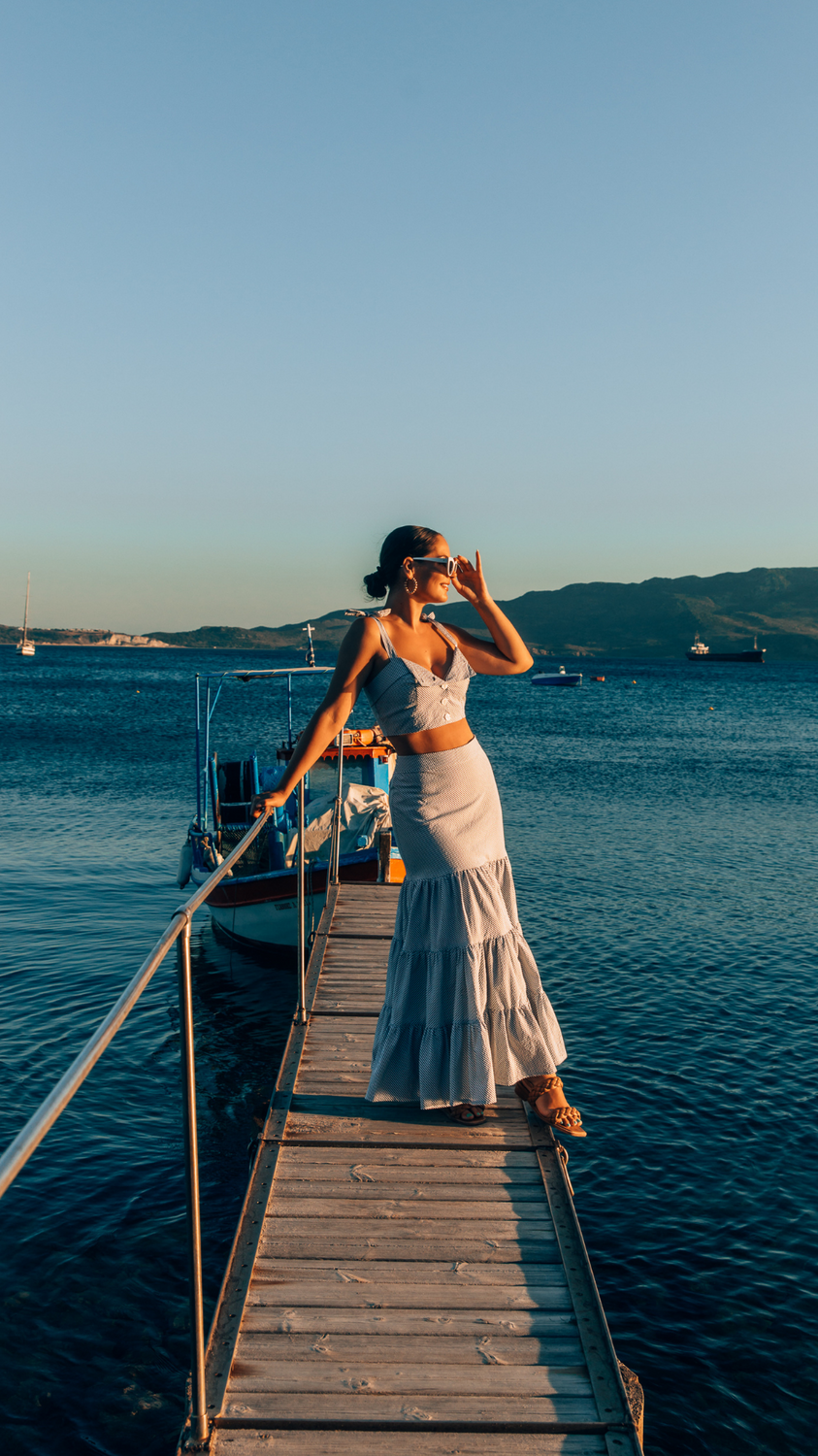  Crop Top and Maxi Skirt set in Blue and White Stripe 100% cotton seersucker deadstock fabric  worn by woman with long brown hair in a bun on a long pier surrounded by blue water, mountains and boats.  She has one hand up to her face touching her sunglasses and looking out towards the sun setting.